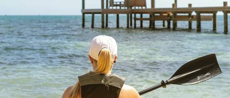 Una persona haciendo kayak en un día despejado, con un muelle de madera y un pabellón al fondo.