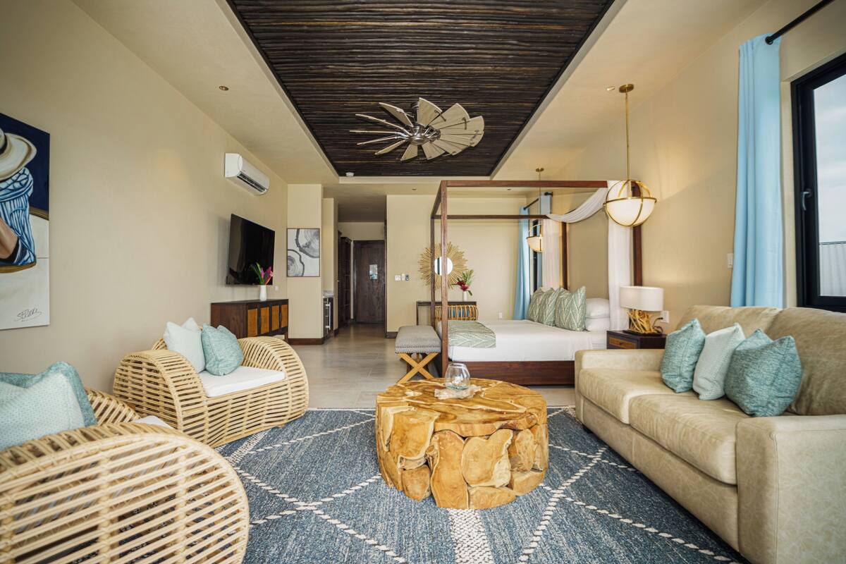 A modern living room with a sofa, wicker chairs, a round coffee table, and a bed in the background.