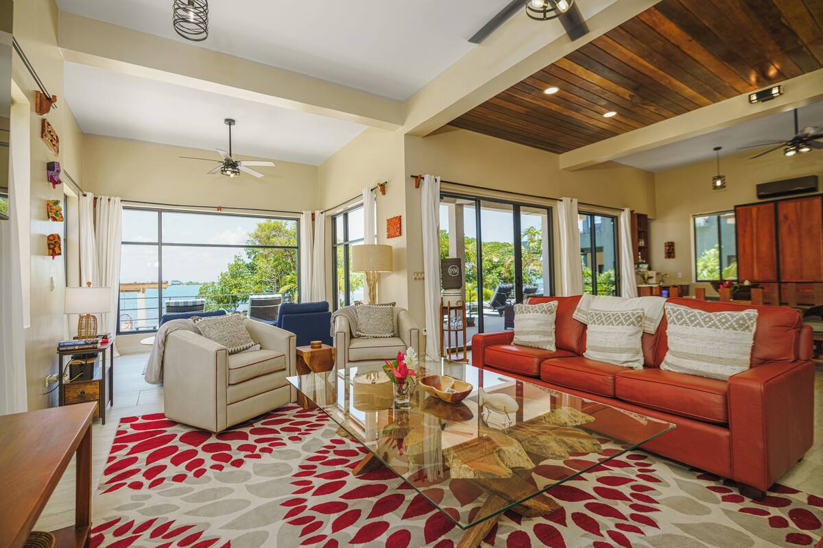 A spacious living room with a large glass coffee table, a red sofa, and a beige sofa. There are large windows providing a view of the outdoors.