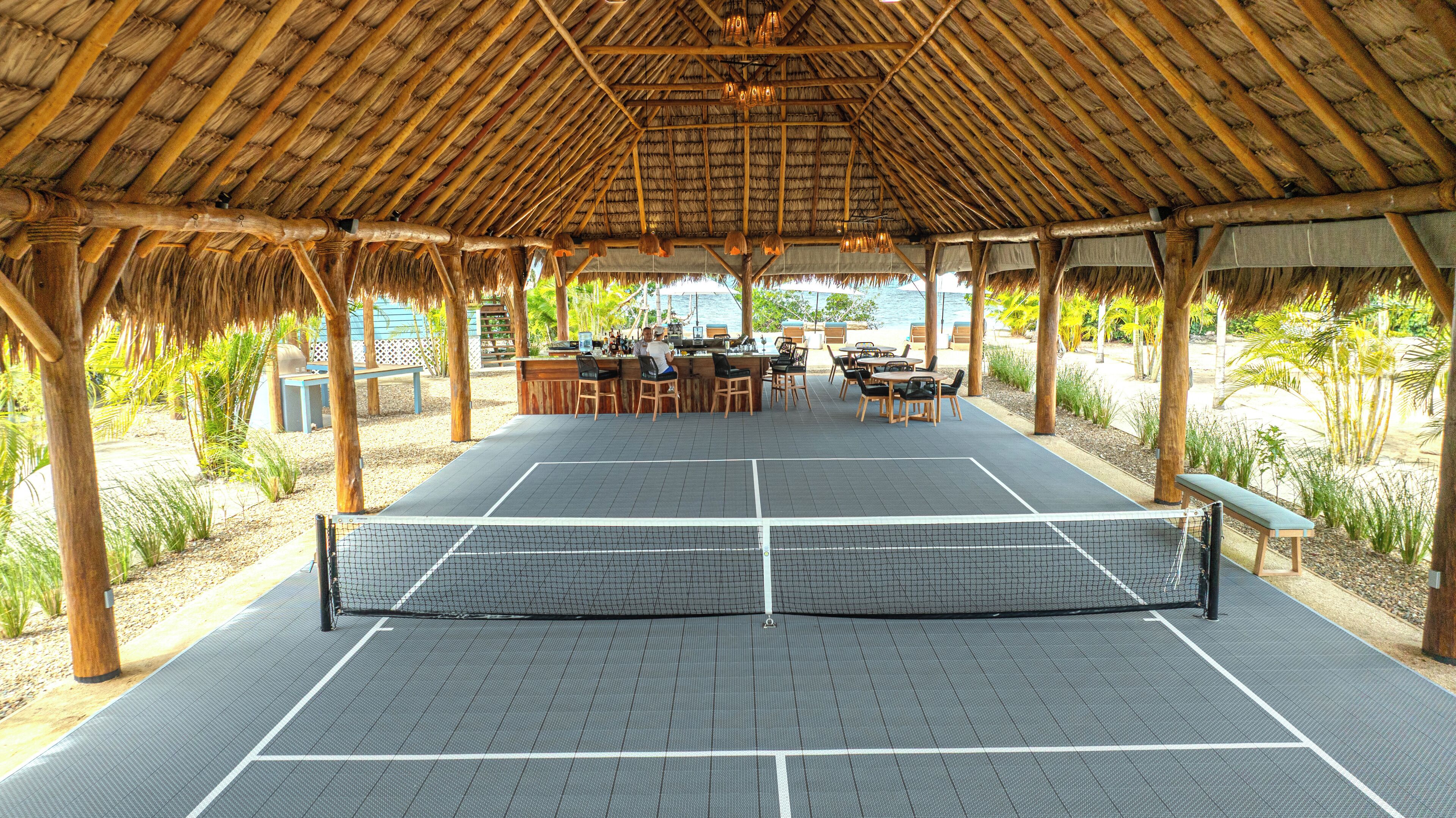 Cancha de tenis exterior con red, rodeada de mesas y sillas de madera, bajo un techo de paja.