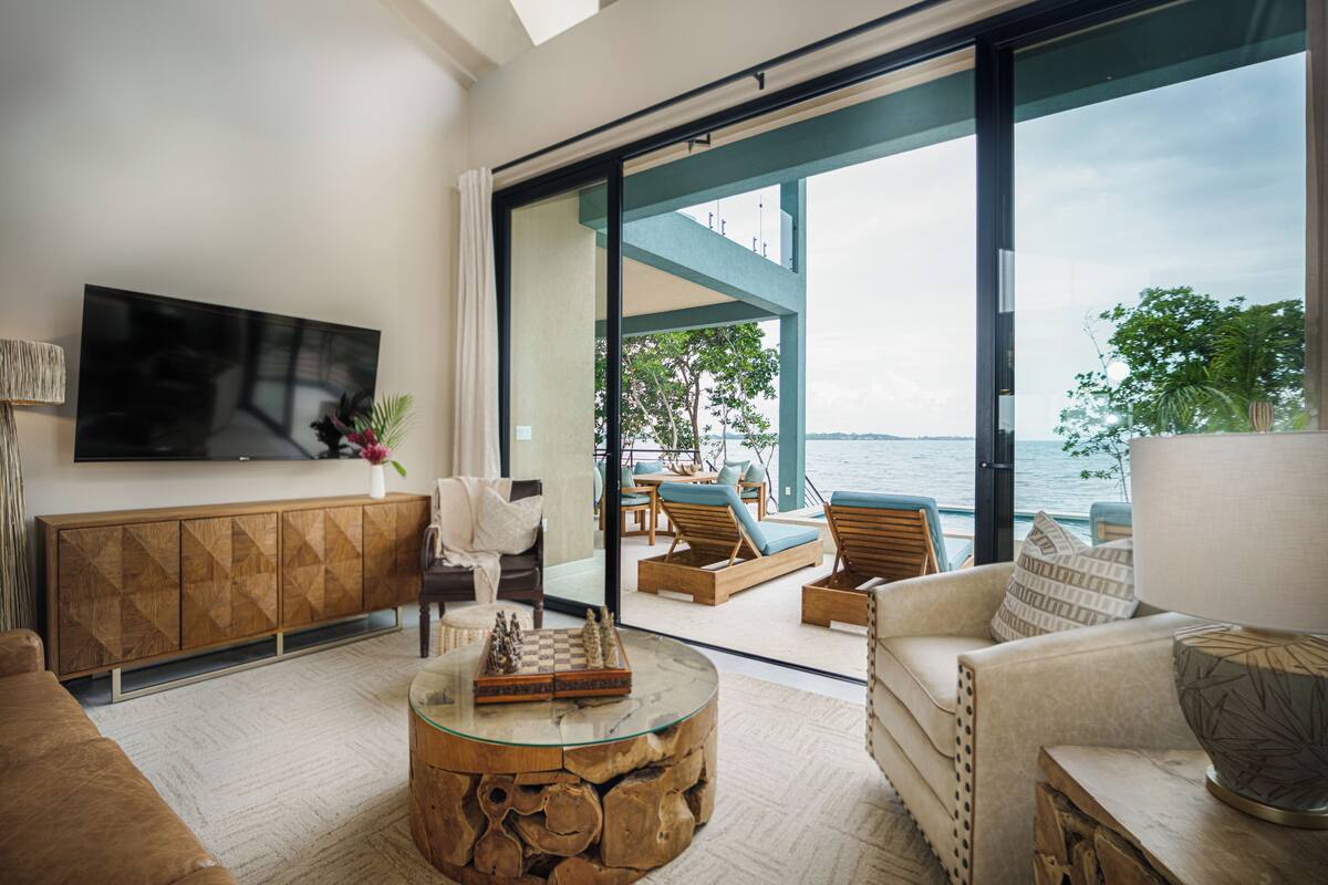 A modern living room with a large TV, comfortable seating, a glass coffee table, and a view of the sea.