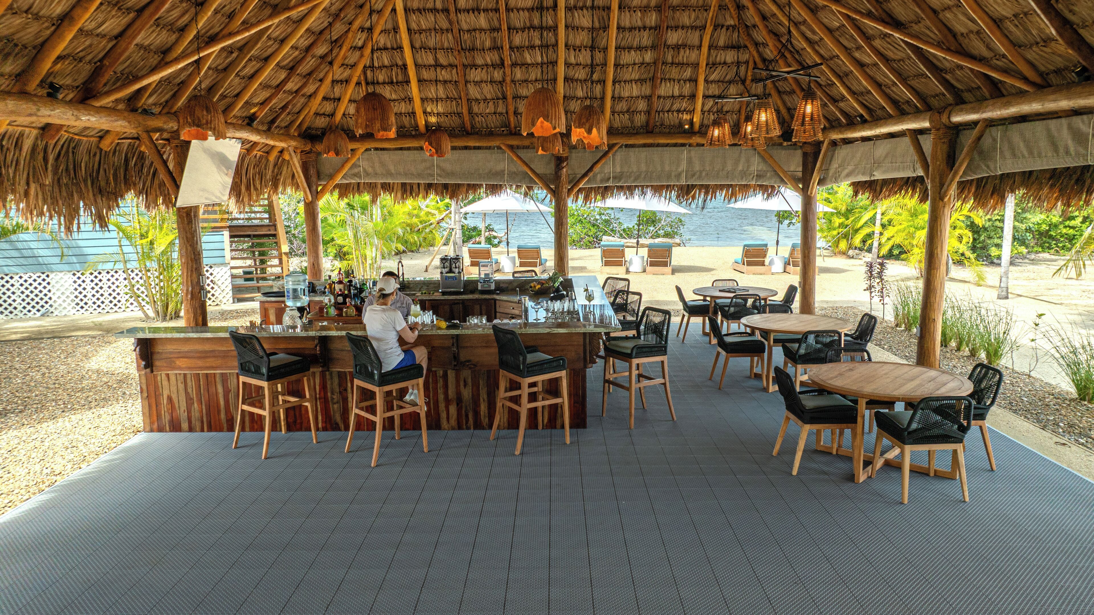 An outdoor bar area with a thatched roof, wooden furniture, and a view of the beach and ocean.