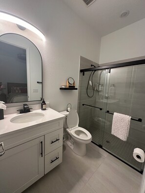Features a sleek vanity with a large mirror and dual sink, alongside a glass-enclosed shower with handheld showerhead and towel rack.