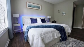 Um quarto com cama grande, mesas de cabeceira, um criado-mudo, uma janela com persianas e uma porta.