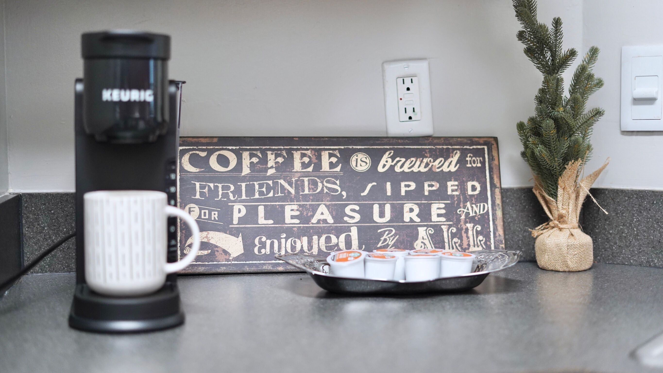 A coffee maker, a mug, a sign with a coffee-related message, a small Christmas tree, and a basket of coffee cups on a kitchen counter.