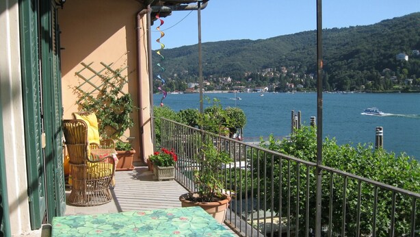 A balcony with a view of a lake, wicker furniture, potted plants, and a table with a patterned tablecloth. - Mansion on Fishermen's Isle (Stresa)