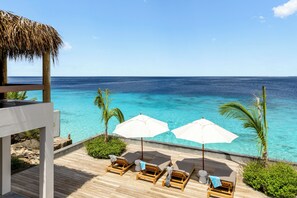 Een houten terras met lounge stoelen en parasols met uitzicht op een heldere blauwe oceaan.