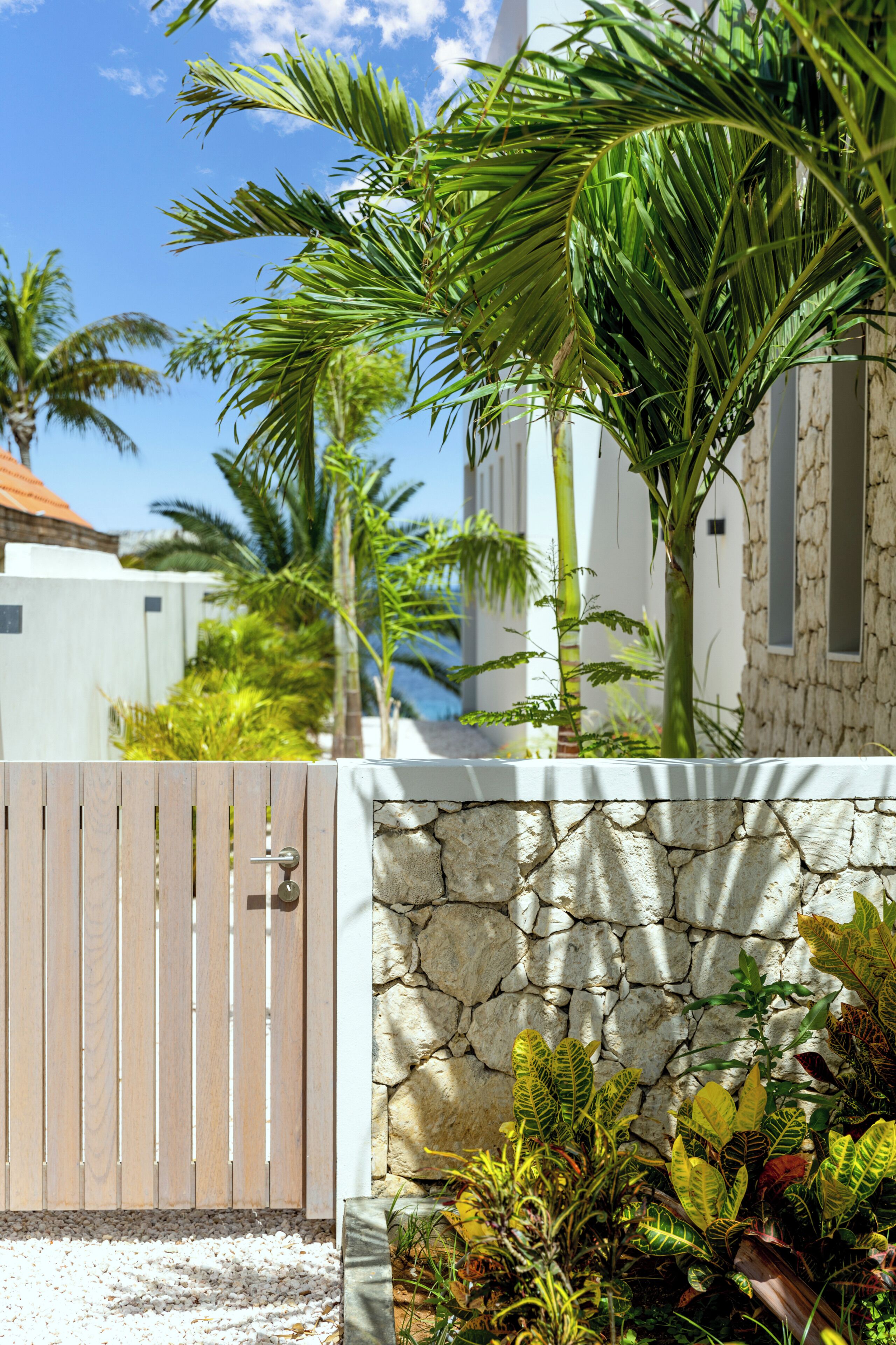 A gated entrance with a wooden fence, a stone wall, and tropical plants.