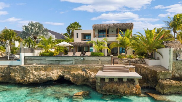 Une propriété située en bord de mer, dotée d’une structure à toit de chaume, d’une eau turquoise cristalline et d’un chemin de pierre menant au bâtiment.