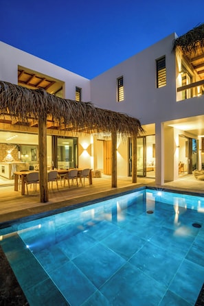 A modern pool area with a thatched roof, a dining area, and a kitchen visible through glass doors.