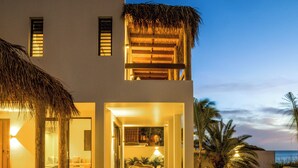 Une maison moderne dotée d’une piscine, d’un balcon surmonté d’un toit de chaume et offrant une vue sur l’océan au crépuscule.