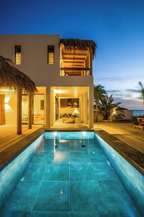 Een modern huis met een zwembad, een balkon met rieten dak en uitzicht op de oceaan bij zonsondergang.