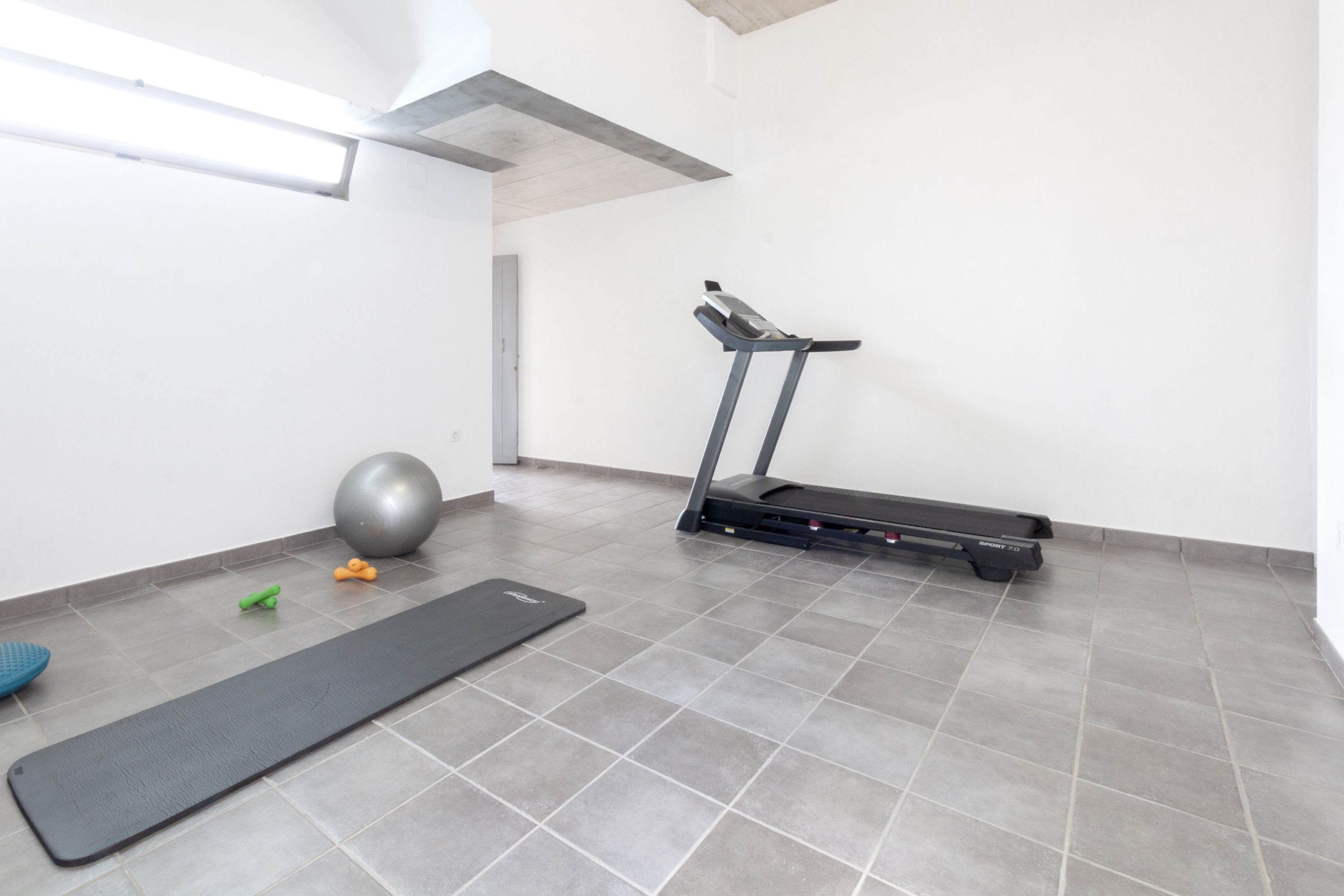 Una habitación pequeña y moderna con suelo de baldosas grises, una colchoneta de ejercicios negra, un balón de ejercicios gris y una cinta de correr.