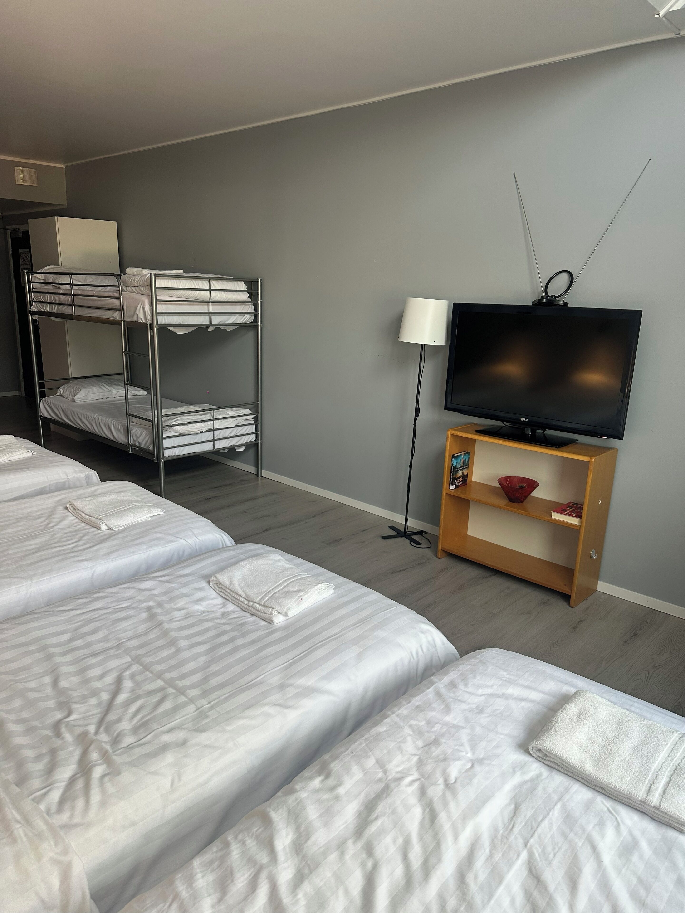 A hotel room with bunk beds, a flat-screen TV, a lamp, and a wooden shelf with a bowl and books.