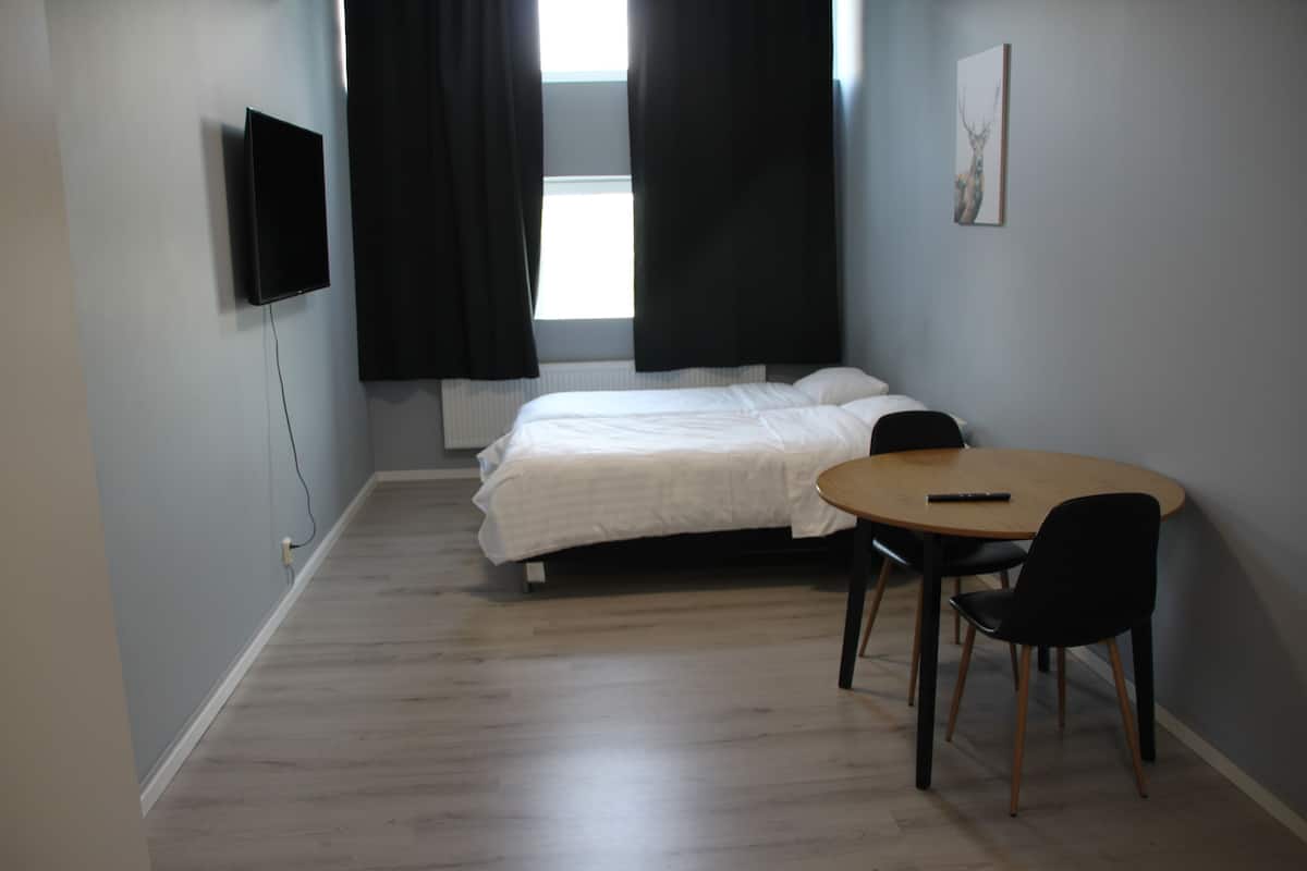 Quarto com cama, televisão fixada na parede, mesa com cadeiras e janela com cortinas.