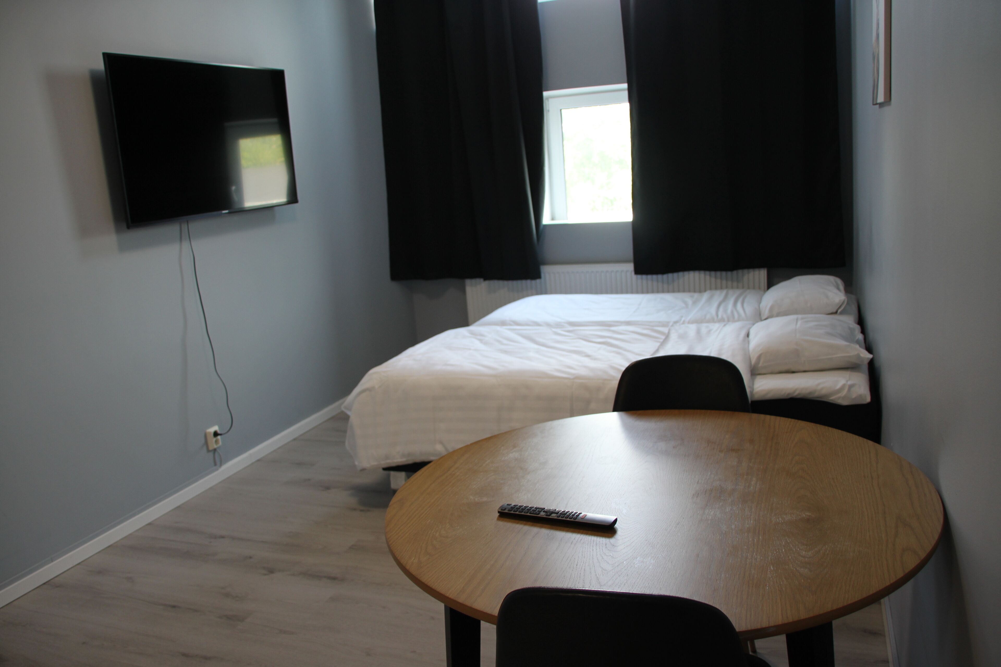 A room with a bed, a TV mounted on the wall, a round wooden table, and a window with curtains.