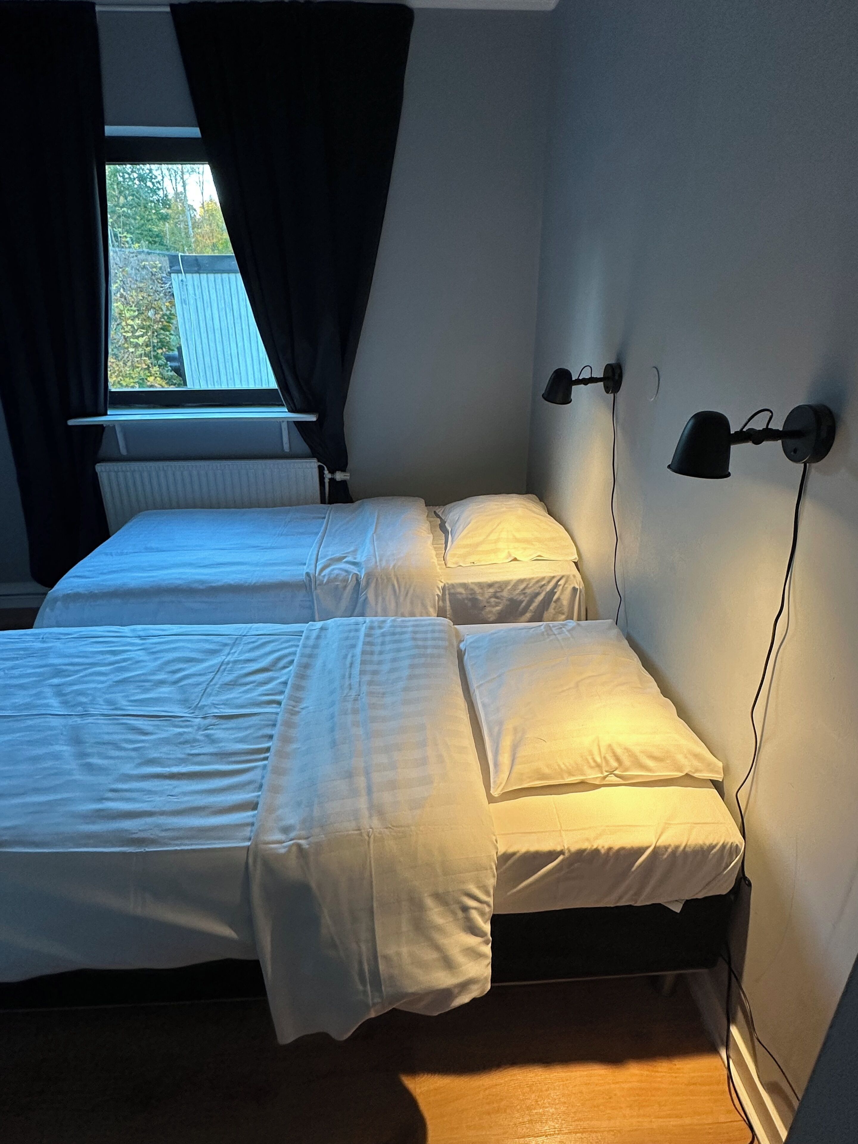 Quarto com duas camas, uma janela com cortinas e luminárias fixadas na parede.