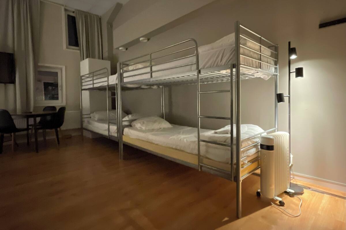 Quarto com beliche, escrivaninha, cadeira, radiador e janela com cortinas.