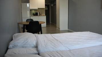 A bedroom with a bed, a small desk, and a chair. There is a kitchenette in the background.