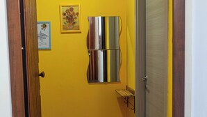 A narrow hallway with yellow walls, a wooden floor, a door with a metal handle, and a small shelf with a framed picture.