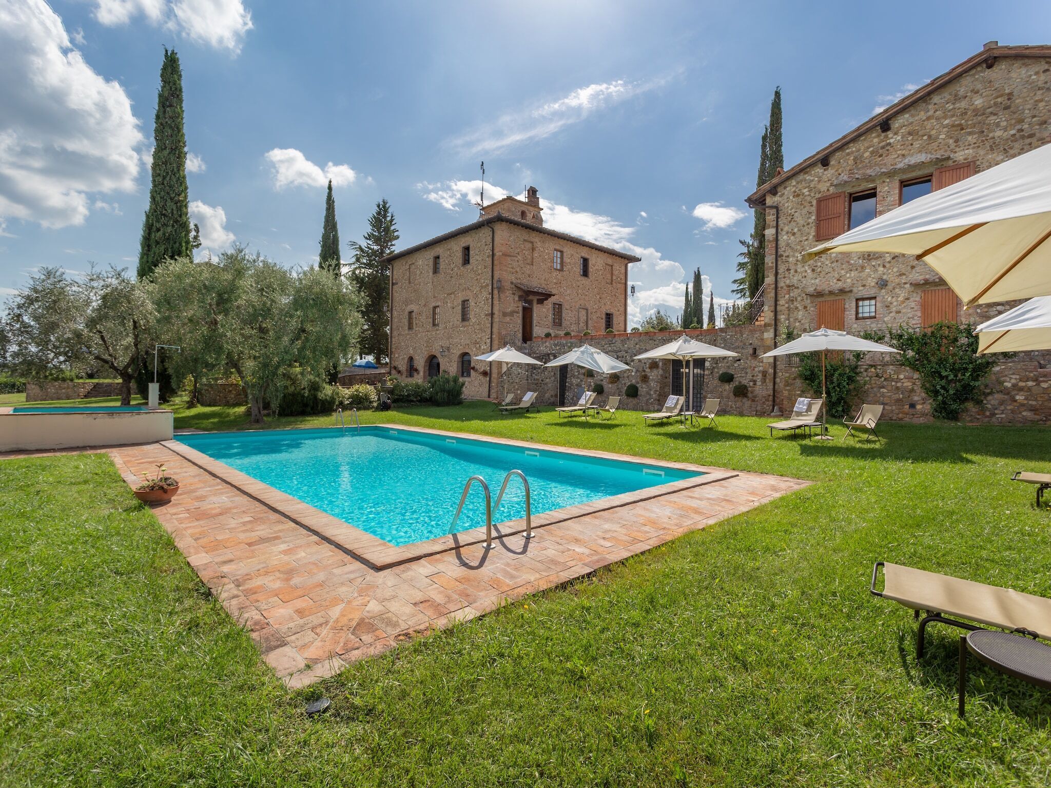 Una piscina con bordo in mattoni, circondata da erba e alberi, con un edificio in pietra sullo sfondo.