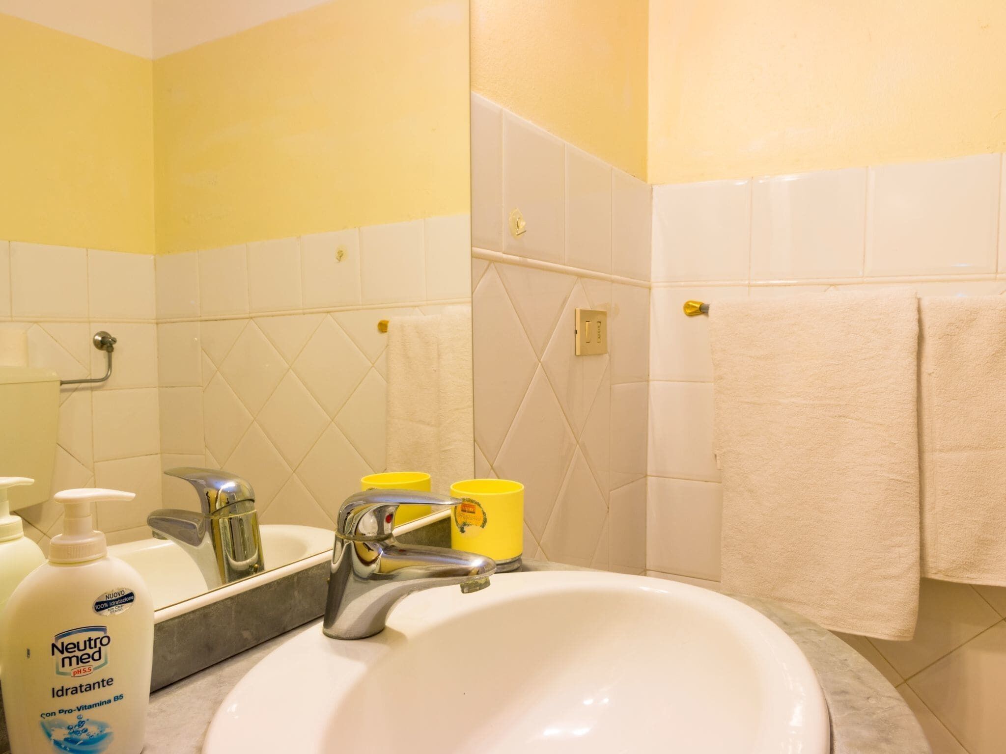 Un baño con lavamanos blanco, paredes con azulejos amarillos y blancos, un espejo y una toalla colgada en la pared.