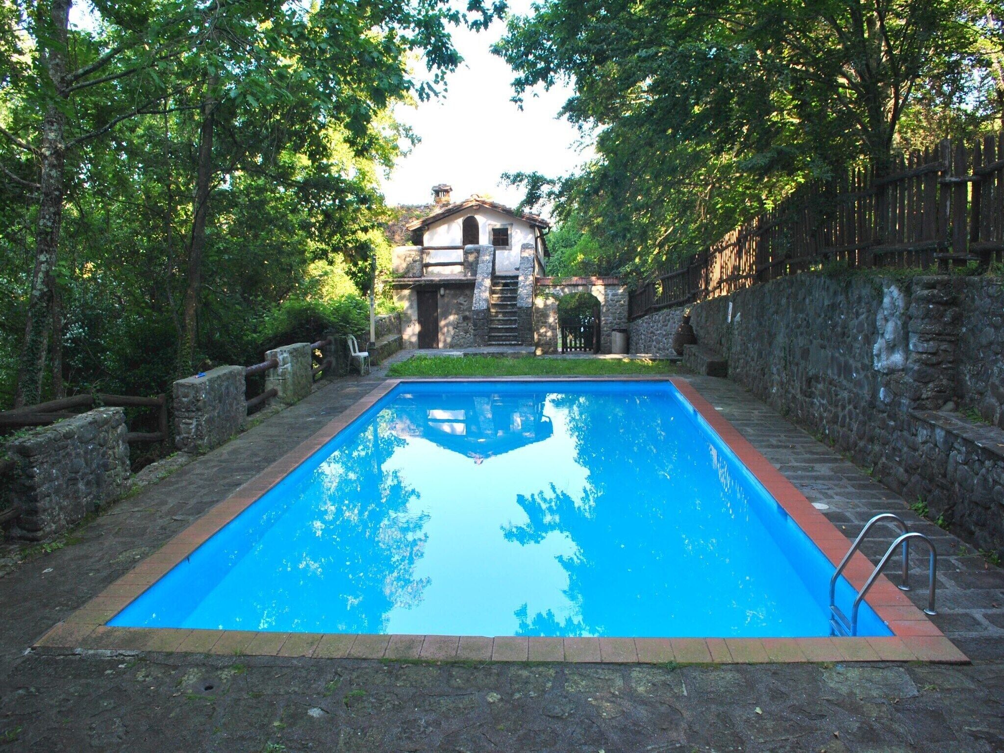 Una piscina rettangolare con bordatura in pietra, circondata da un muro in pietra e un recinto in legno.
