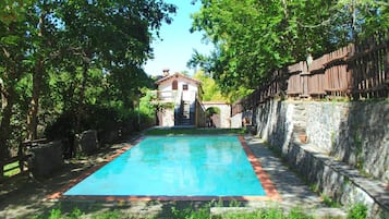 Una piscina con un muro in pietra da un lato e una recinzione in legno dall'altro, che conduce a un edificio con tetto in tegole rosse.
