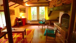 Ein gemütliches Wohnzimmer mit Kamin, Holzbalken, einem Esstisch und Blick durch ein Fenster mit gelben Vorhängen.