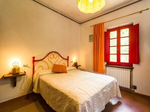 Un dormitori amb llit, una tauleta de nit, una làmpada, una finestra amb cortines i un radiador.