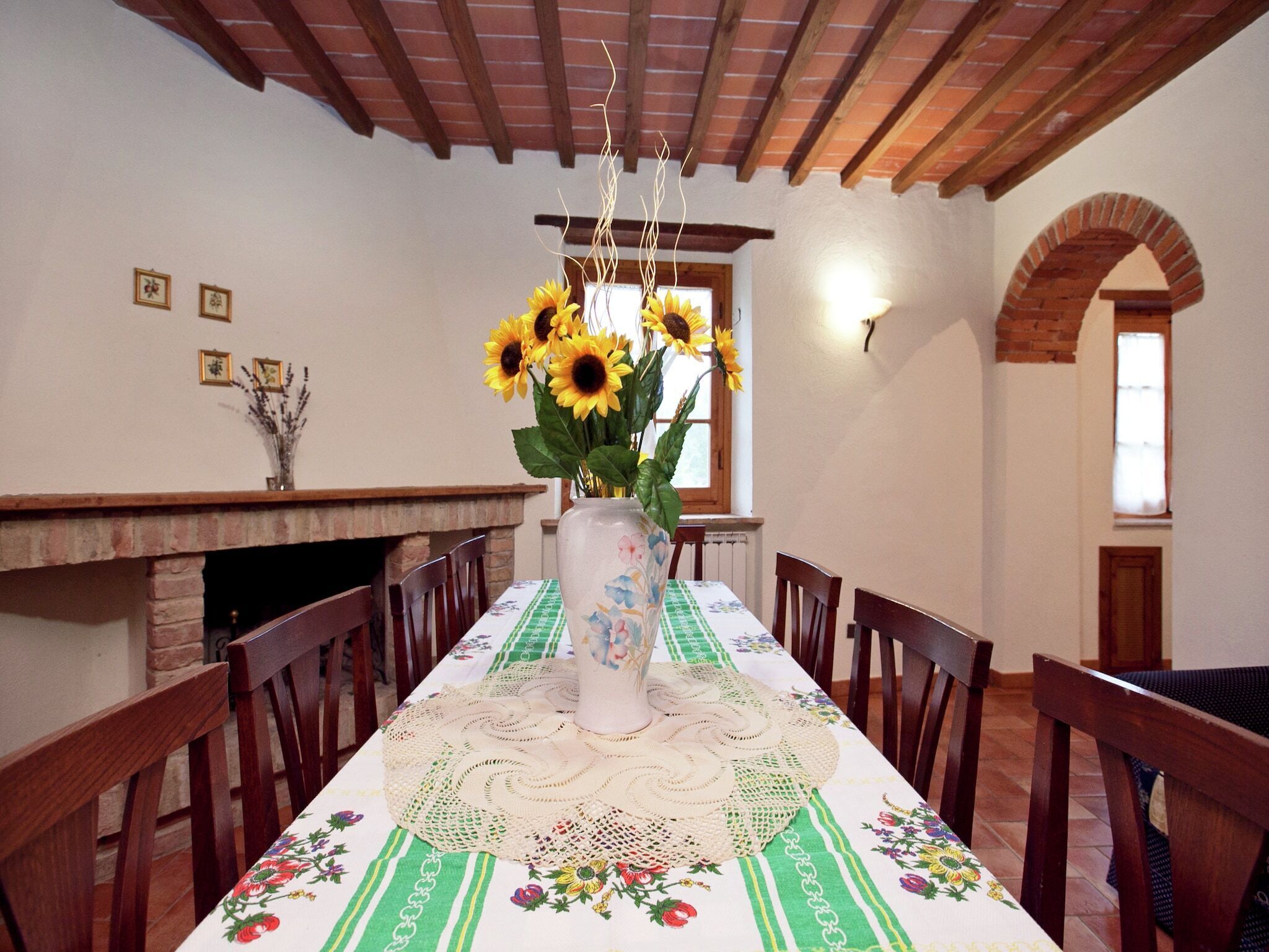 Une salle à manger avec une longue table, des chaises, un vase rempli de tournesols, une cheminée en briques et une fenêtre donnant sur un beau paysage.