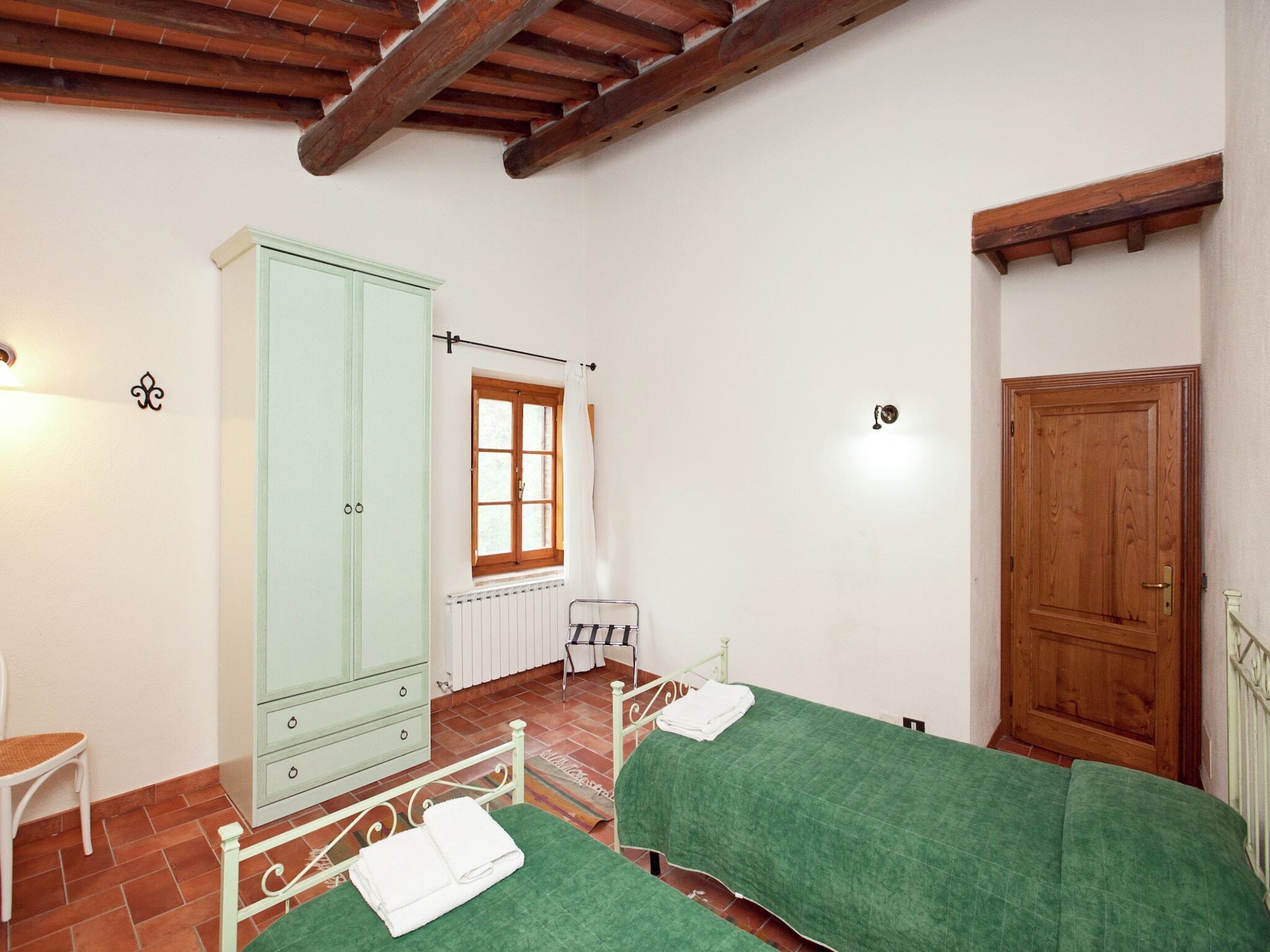 A room with two beds, a radiator, a window with curtains, a wooden door, and a cabinet.