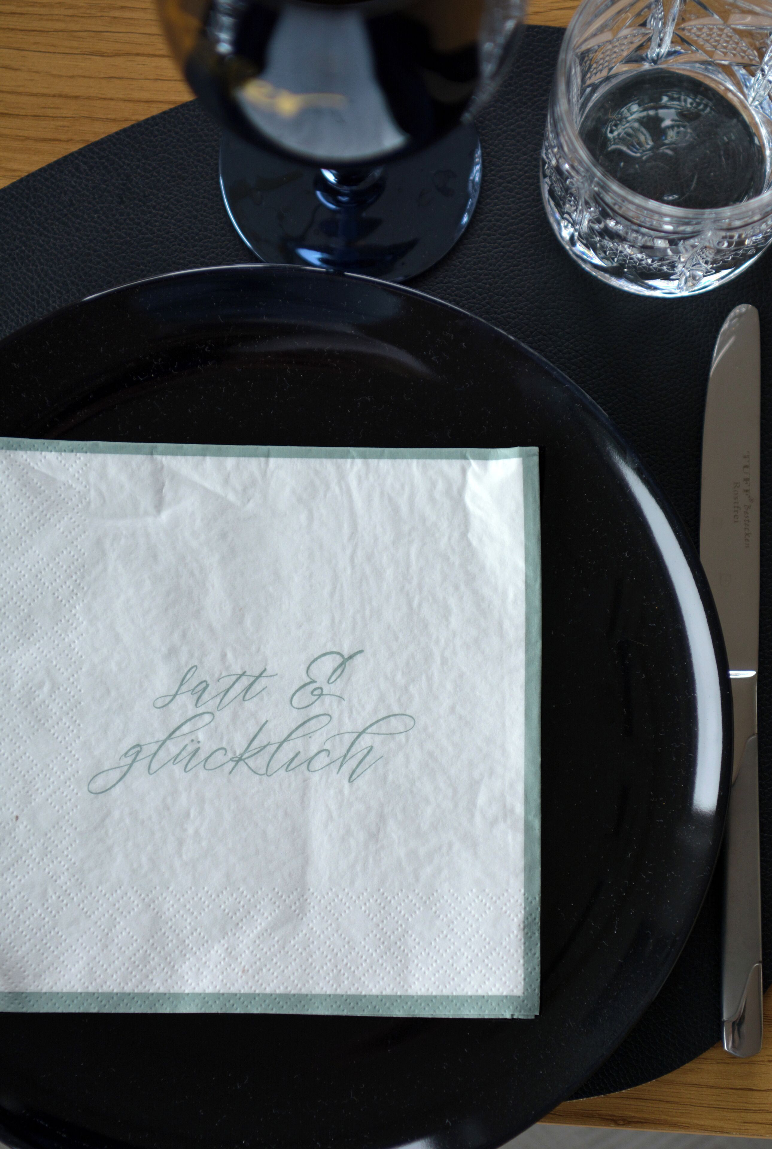 Une assiette noire avec une serviette, un couteau et un verre d’eau, le tout posé sur une table.
