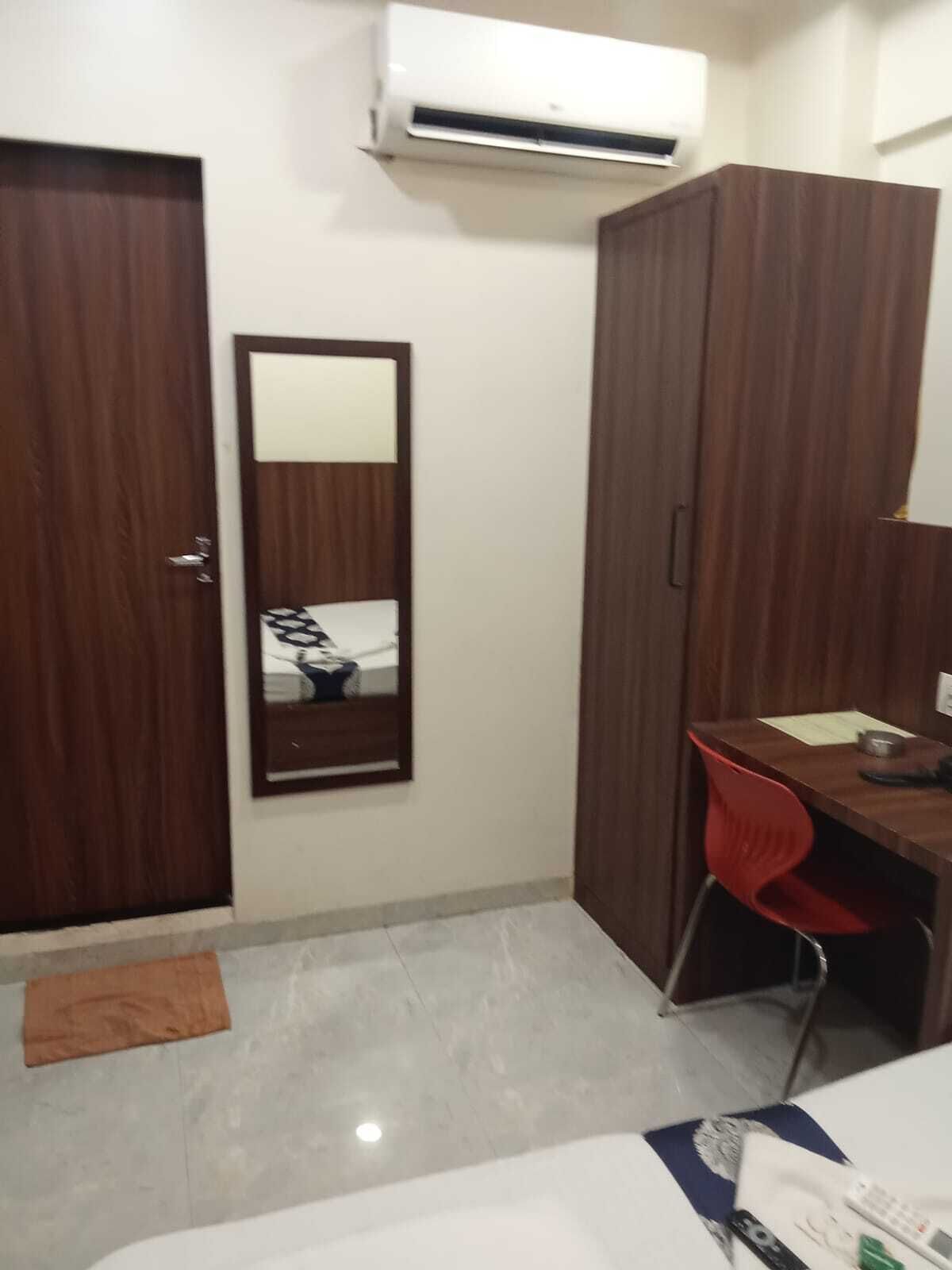 A small, neatly arranged bedroom with a wooden wardrobe, a desk with a chair, a bed, and an air conditioning unit mounted on the wall.
