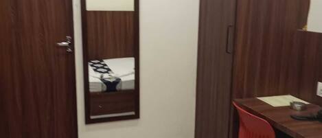A small, neatly arranged bedroom with a wooden wardrobe, a desk with a chair, a bed, and an air conditioning unit mounted on the wall.
