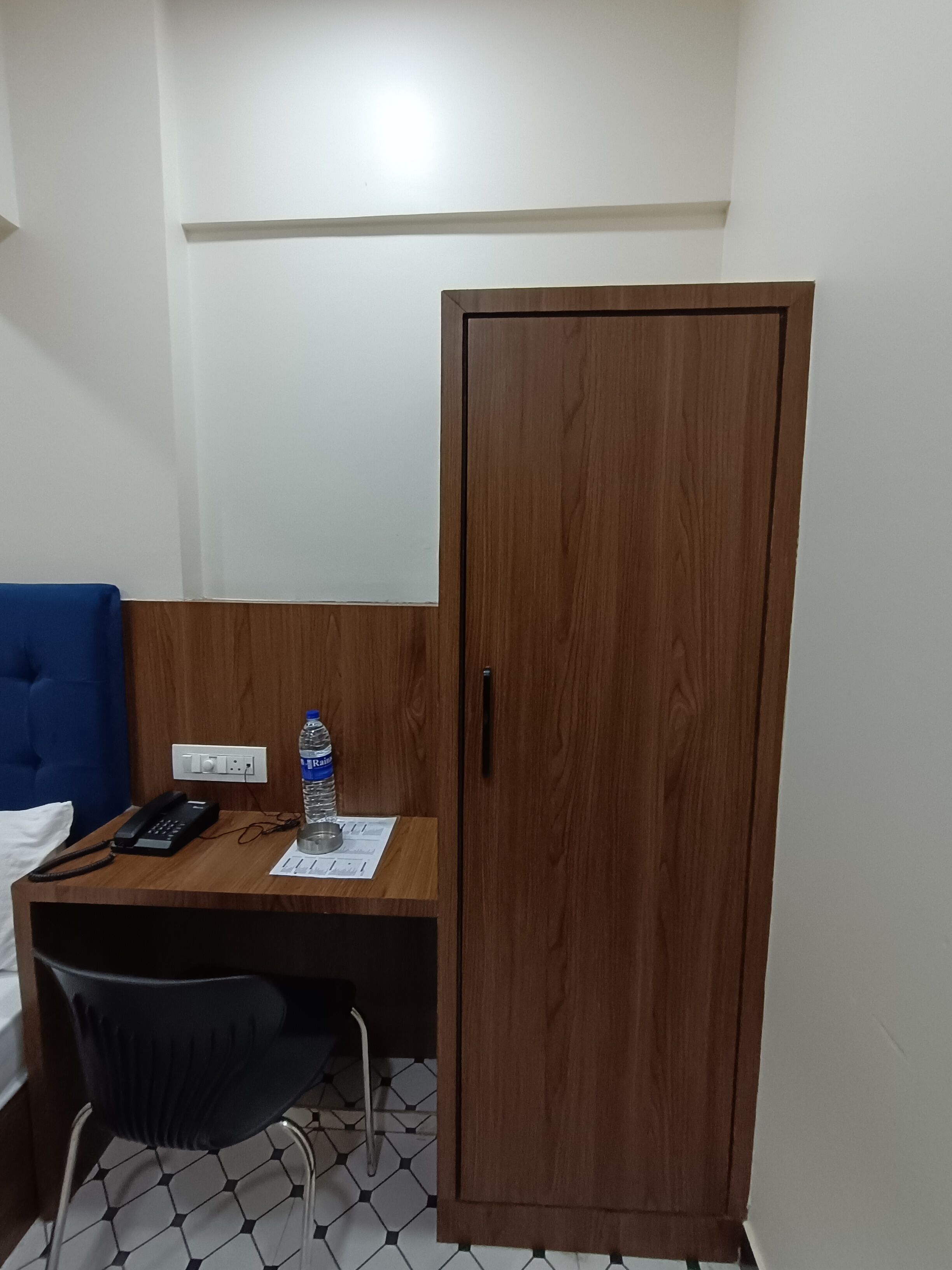 A small hotel room with a wooden desk, a blue chair, a wardrobe, a telephone, and a water bottle.