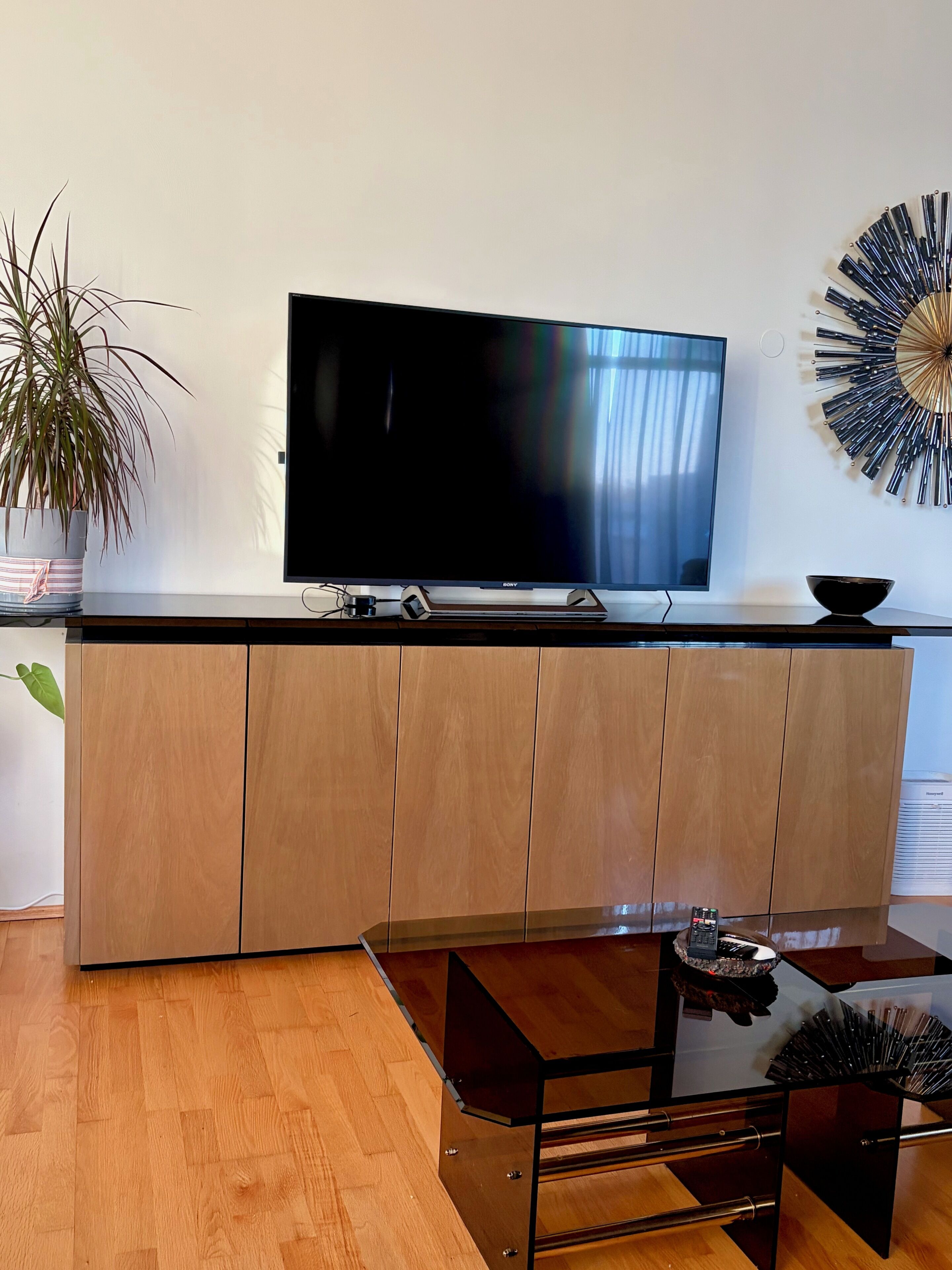 A modern living room with a flat-screen TV, wooden cabinets, a glass coffee table, and a decorative wall piece.