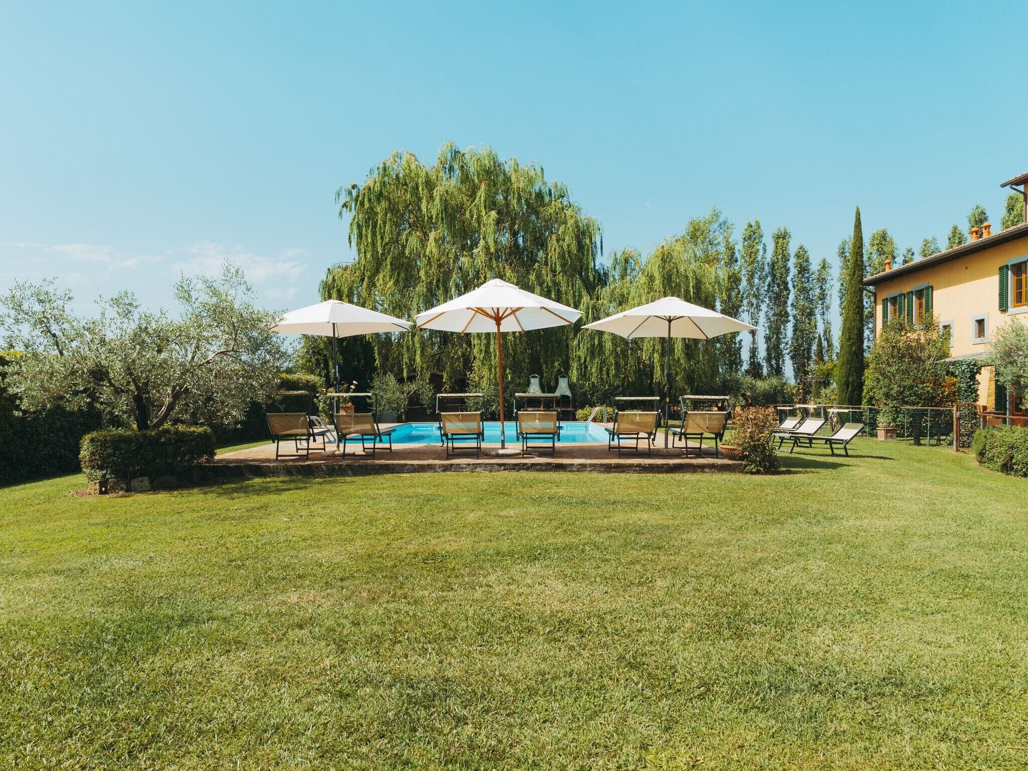 A pool area with lounge chairs and umbrellas, surrounded by greenery and a building in the background.
