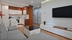 Un salon moderne avec un téléviseur à écran plat fixé sur un mur en briques blanches, un canapé, une table basse et un coin cuisine à l’arrière-plan.