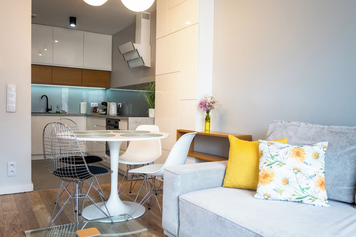Une cuisine moderne avec des meubles blancs, un coin repas doté d’une table ronde et de chaises, et un salon avec un canapé et des coussins.