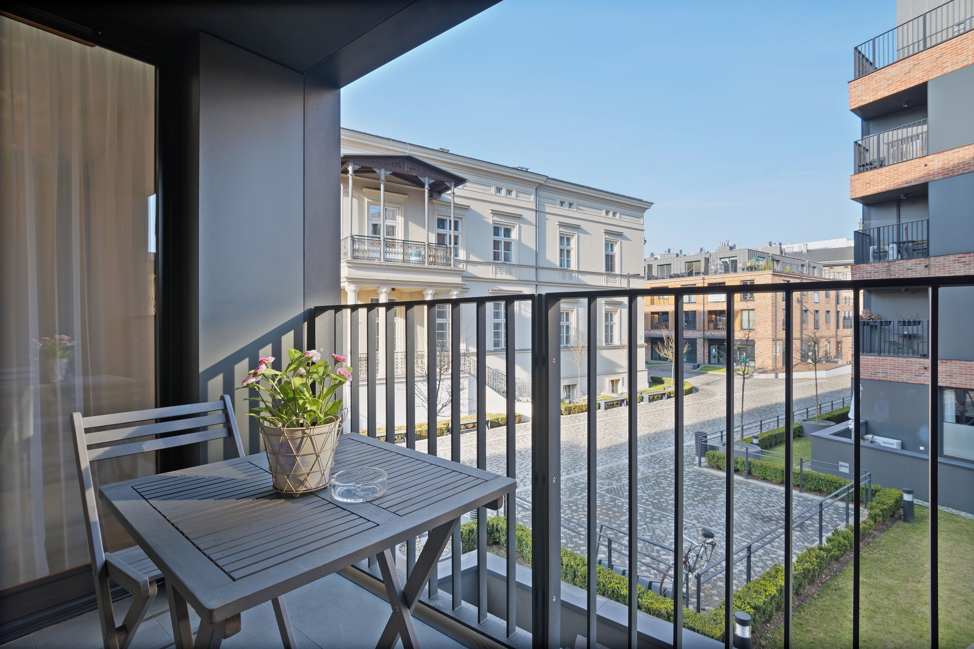 A balcony with a table and chair, a potted plant, and a view of a street with buildings.