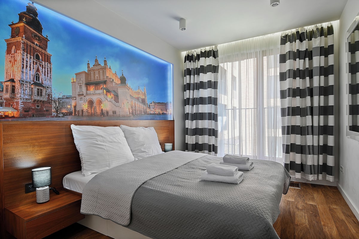Une chambre d’hôtel avec un grand lit, une table de chevet avec une lampe, une fresque murale représentant un bâtiment historique, et des rideaux à rayures noires et blanches.