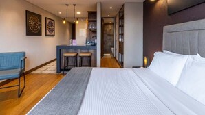 A modern hotel room with a large bed, a desk with two chairs, a blue sofa, and a small kitchenette.