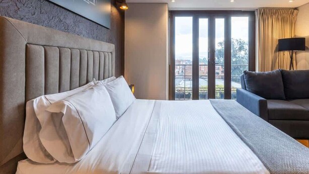 A modern hotel room with a large bed, a sofa, and a view of the city through large windows.