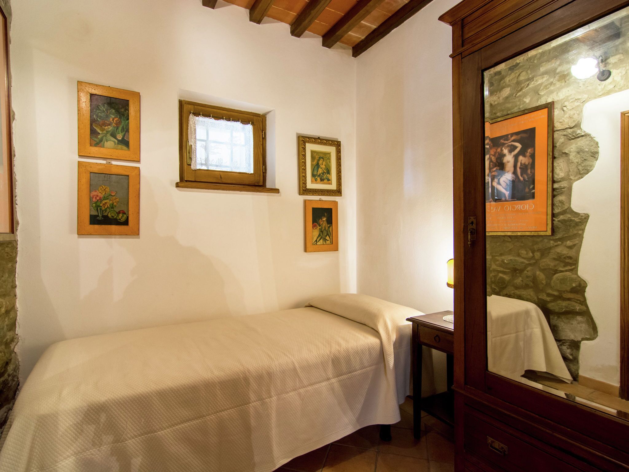 Une petite chambre avec un lit simple, une tête de lit en bois, une petite table avec une lampe et plusieurs tableaux encadrés accrochés aux murs.