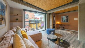 Una sala de estar moderna con sofá, reposapiés, mesa de centro y vistas a edificios a través de amplios ventanales.