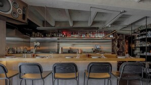 Una zona de bar con mostrador de acero inoxidable, taburetes y una variedad de botellas en estantes.
