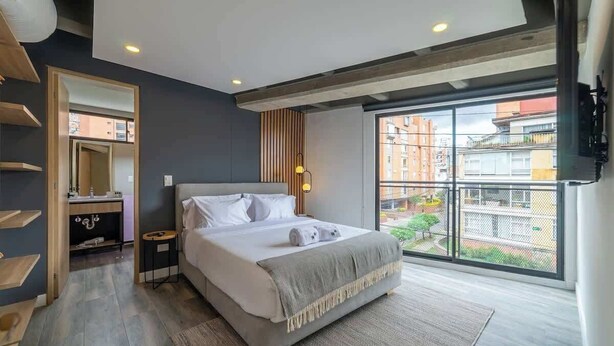 Un dormitorio moderno con una cama grande, una mesita de noche, un espejo y una ventana con vistas a edificios.