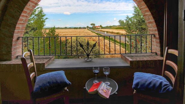 Een balkon met uitzicht op een veld en een huis in de verte, met twee stoelen en een tafel met glazen en een boek.
