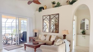 A spacious living room with a large sofa, a coffee table, and a ceiling fan. There is a glass door leading to an outdoor area with a patio table and chairs. - CasaSuLago Sunset Lakefront, Saltwater Pool & Spa, Fishing, Kayaks & Game Room! (Cape Coral)